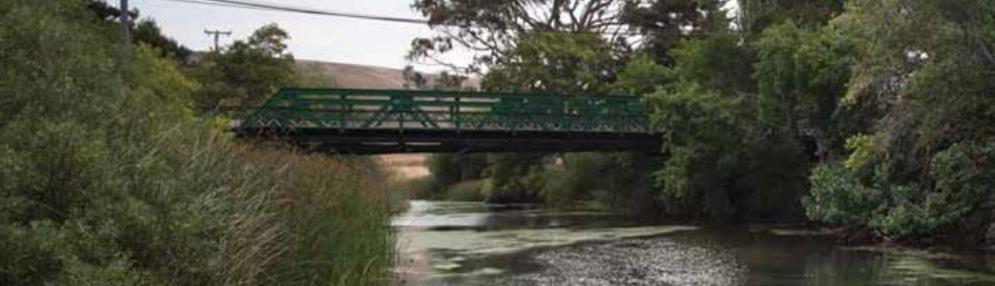 Photograph of the Lagunitas Creek Bridge spanning Lagunitas Creek near Point Reyes Station in Marin County. The green colored bridge connects the tree-lined shores on either side of the stream that splits the middle of the image.